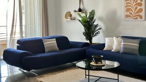 Un salon moderne avec un canapé bleu, une table basse ronde, un luminaire suspendu, une plante en pot et une œuvre d’art murale.