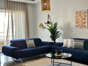 A modern living room with a blue sofa, a round coffee table, a hanging light fixture, a potted plant, and a wall art piece.