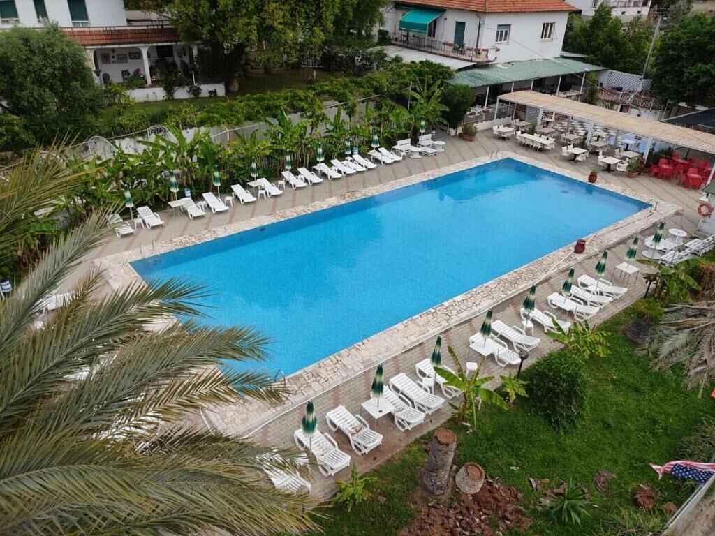 A rectangular swimming pool with lounge chairs and umbrellas, surrounded by greenery and buildings.