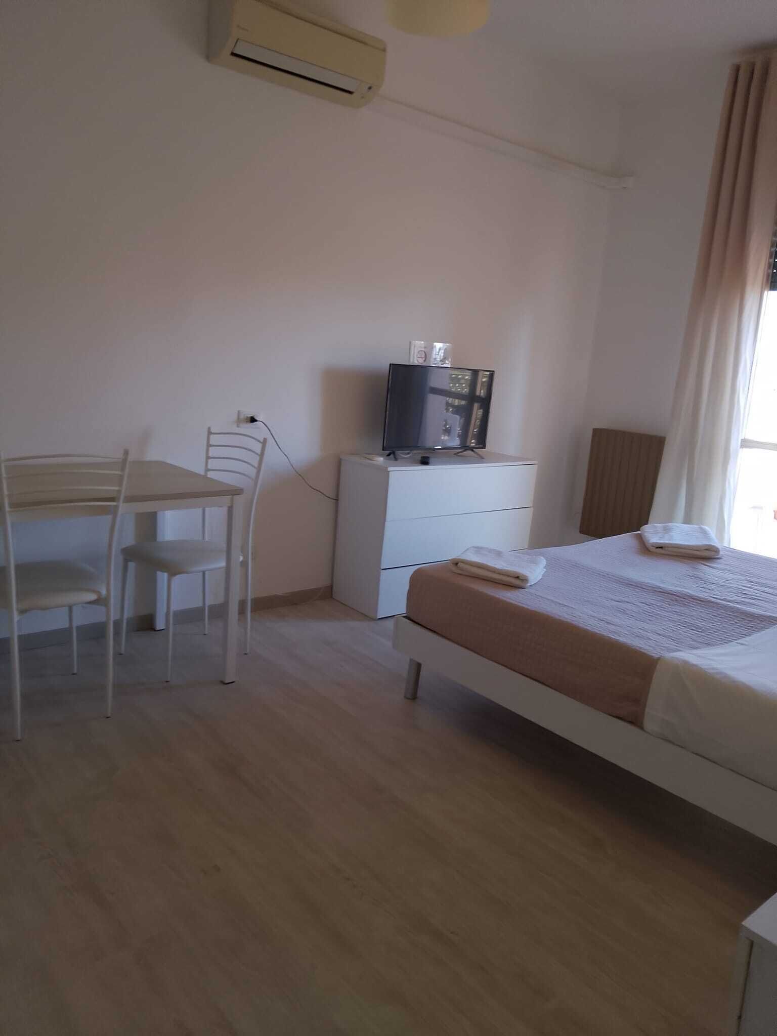 A bedroom with a bed, a desk with a chair, a TV on a dresser, and an air conditioning unit.
