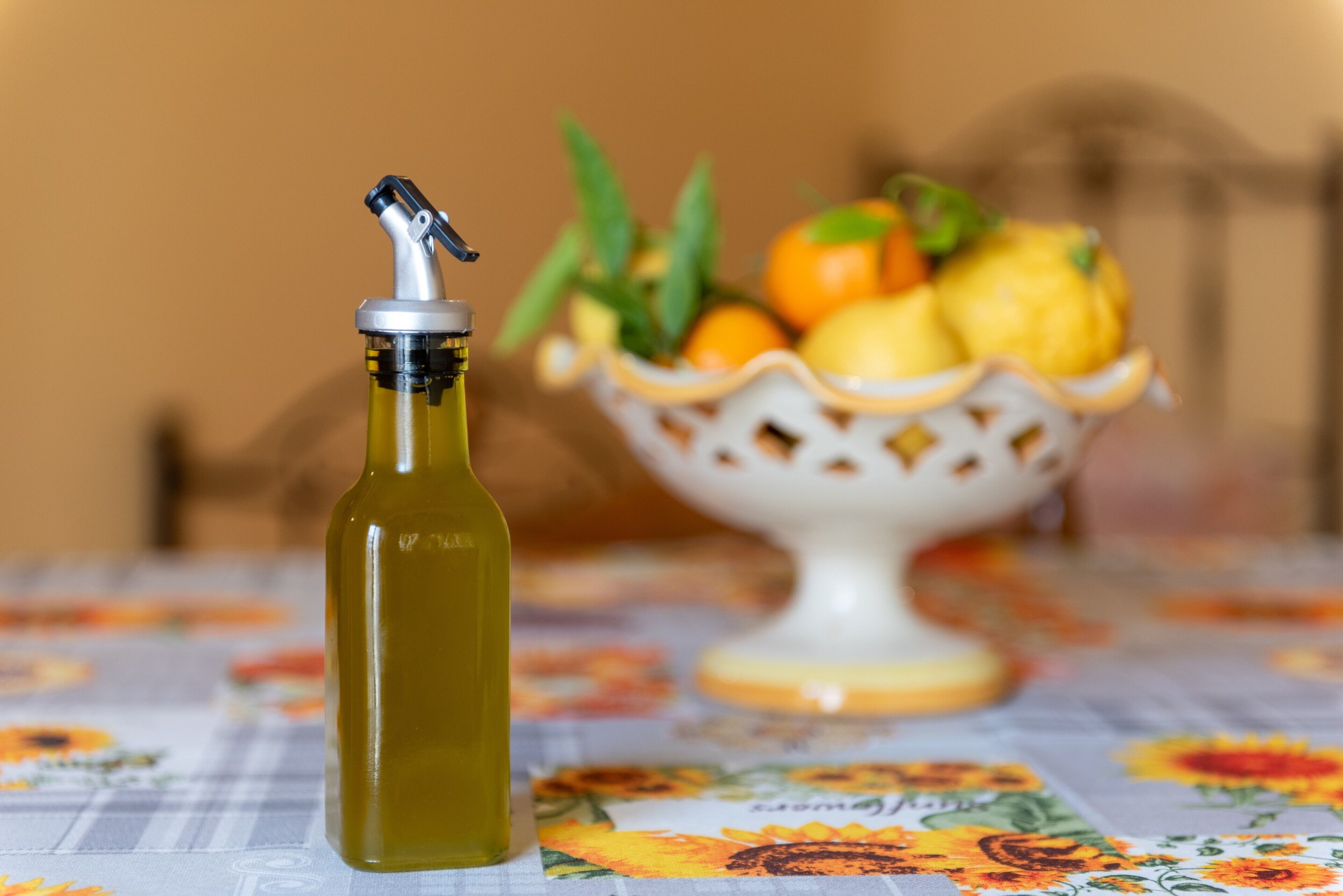 Une bouteille d’huile d’olive est posée sur une table recouverte d’une nappe à motifs floraux, et un saladier de fruits se trouve à l’arrière-plan.