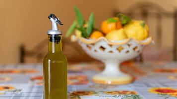 Une bouteille d’huile d’olive est posée sur une table recouverte d’une nappe à motifs floraux, et un saladier de fruits se trouve à l’arrière-plan.