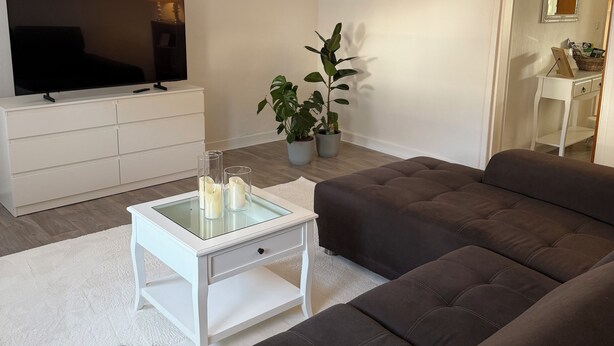 Una sala de estar moderna con un televisor de pantalla plana, una mesa de centro blanca, un sofá gris y un otoman blanco y mullido.