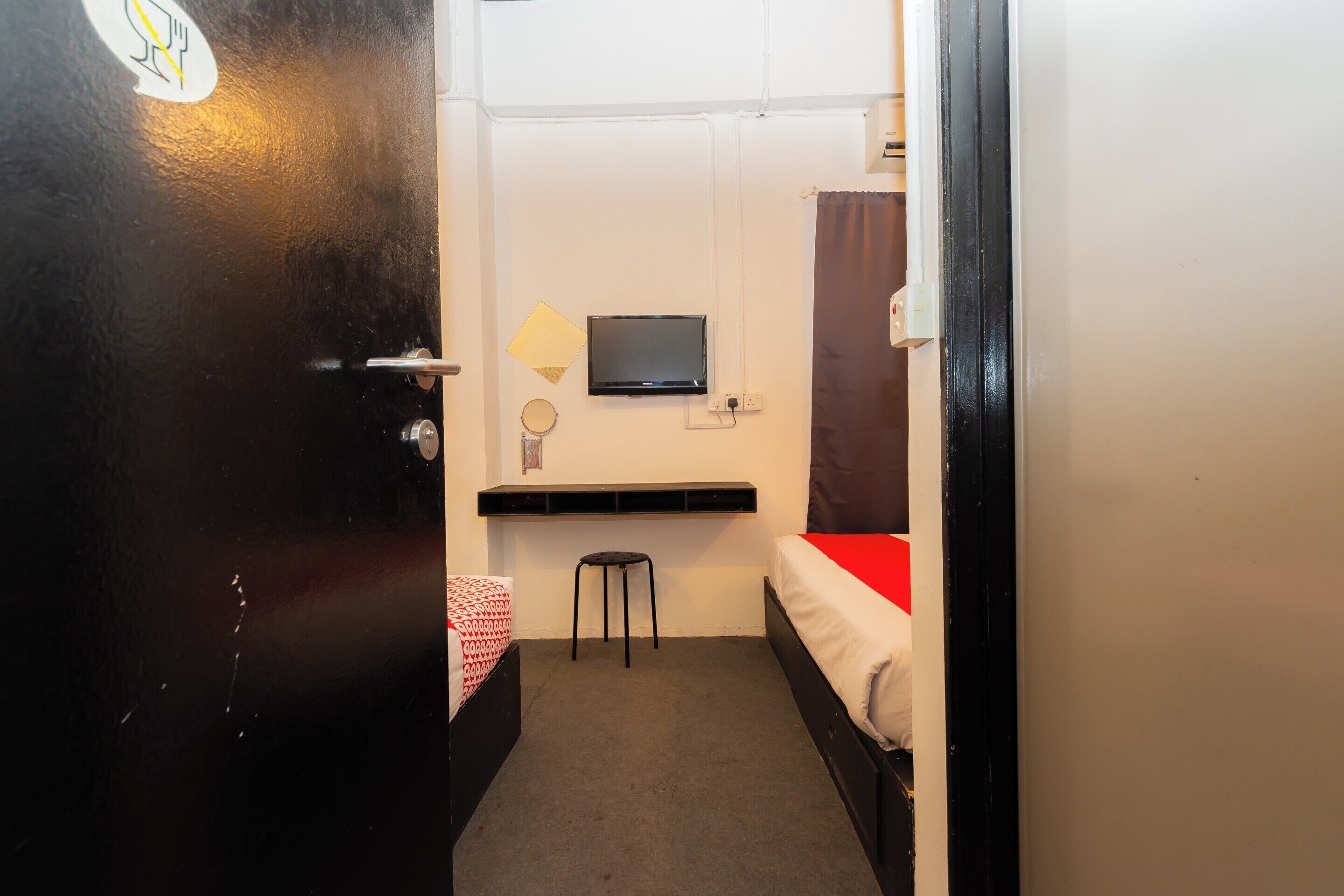 A compact room with a single bed, a small stool, a TV mounted on the wall, and a shelf above the bed.