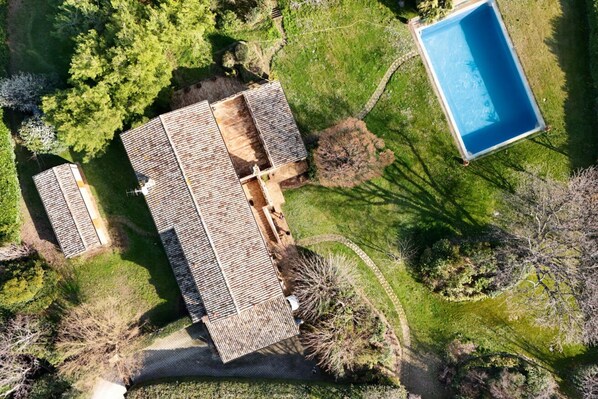 An aerial view of a property with a swimming pool, a large building with a sloped roof, and surrounding greenery.