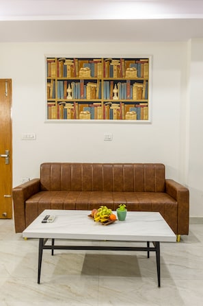 A room with a brown leather sofa, a white table with fruits and a remote control, and a bookshelf with books and decorative items. - The Empire Sector 45 Gurgaon (Gurugram)