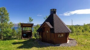 Una cabaña de madera con techo cónico, una estructura de madera separada y vistas a un lago y montañas.