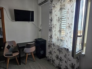 Ein Zimmer mit einem wandmontierten Flachbildfernseher, einem Schreibtisch mit Stuhl, einer Klimaanlage und einem Fenster mit blumigen Vorhängen.