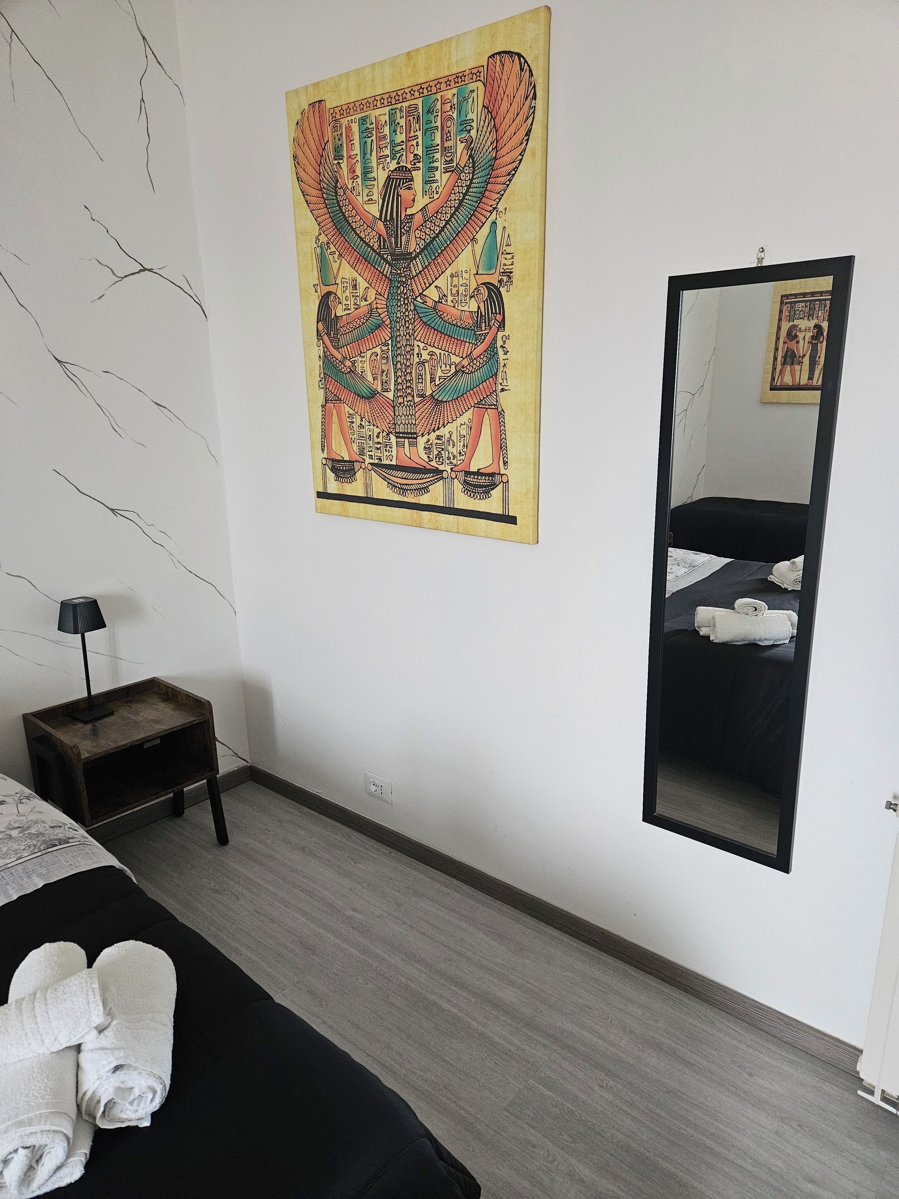 Ein Hotelzimmer mit einem Bett, einem kleinen Nachttisch mit Lampe, einem Wandspiegel und einem Wandbild.