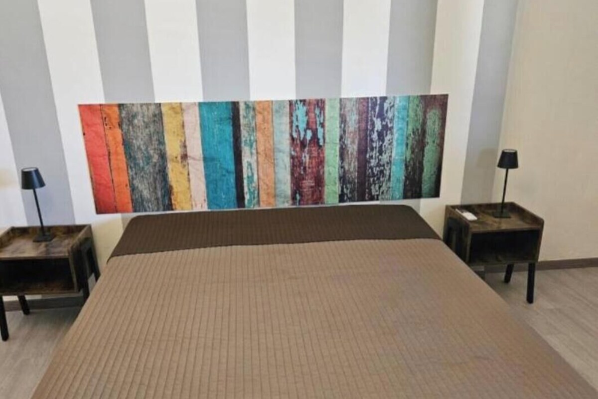A hotel room with a large bed, two nightstands, a colorful abstract painting on the wall, and a striped pattern on the ceiling.