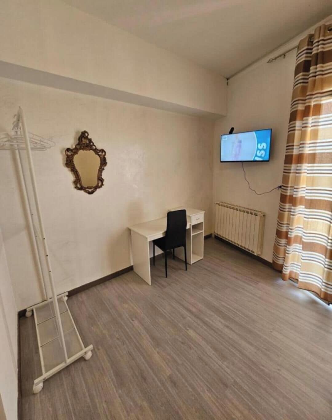 Quarto com escrivaninha, cadeira, televisão, radiador e janela com cortinas.