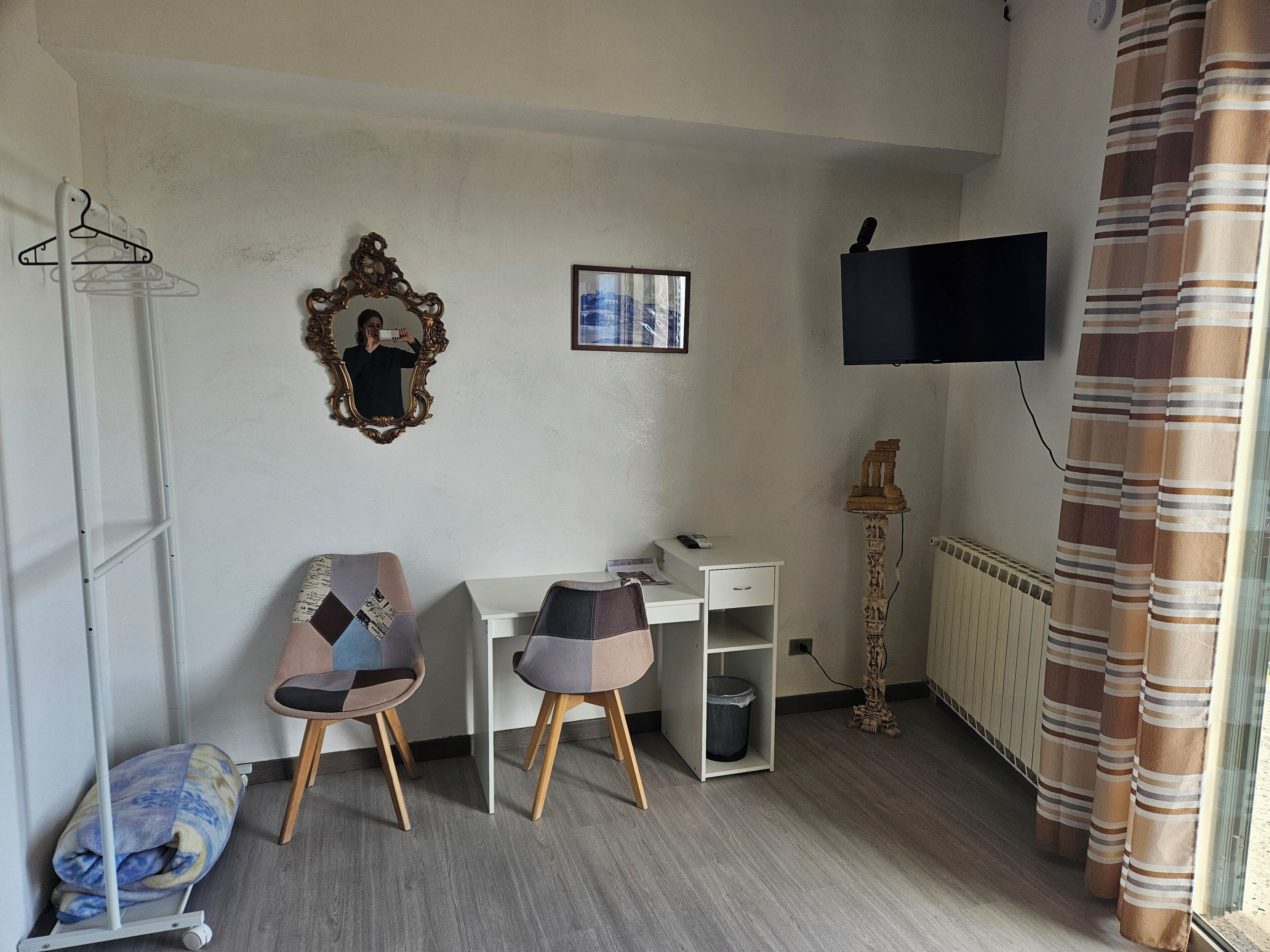 Ein Zimmer mit Schreibtisch, zwei Stühlen, einem Spiegel, einem Fernseher, einer Heizung und einem Vorhang.