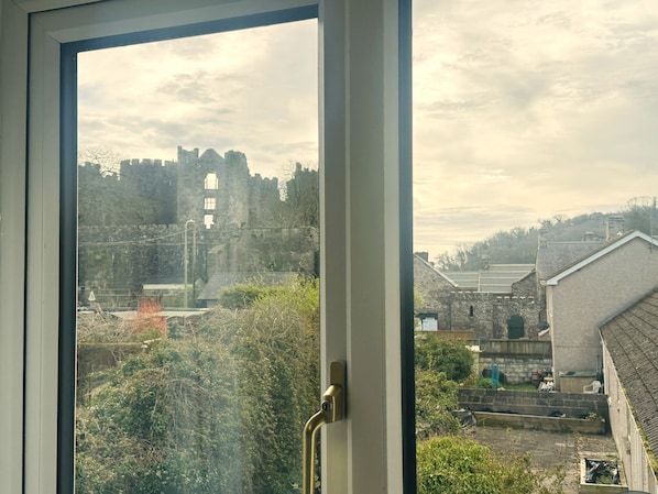 A view through a window with a metal handle, a stone building with a tower, and a cloudy sky.