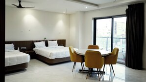 A hotel room with two beds, a dining table with chairs, a ceiling fan, and a large window with curtains. - HOTEL LEELA INN (Asnawar)