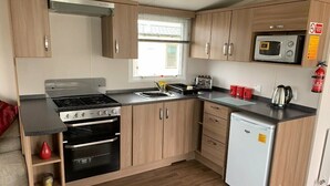 A compact kitchen with wooden cabinets, a microwave, a stove, and a small refrigerator. - Berwick Holiday Park - Holiday Accommodation 4088 (Berwick-upon-Tweed)