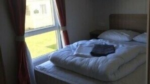 A single bed with a wooden headboard, a bedside table, and a window with curtains. - Berwick Holiday Park - Holiday Accommodation 4088 (Berwick-upon-Tweed)