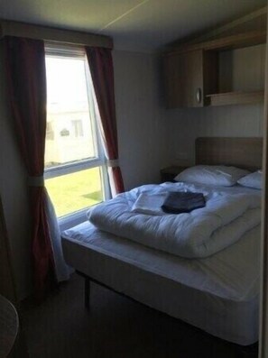 A single bed with a wooden headboard, a bedside table, and a window with curtains. - Berwick Holiday Park - Holiday Accommodation 4088 (Berwick-upon-Tweed)