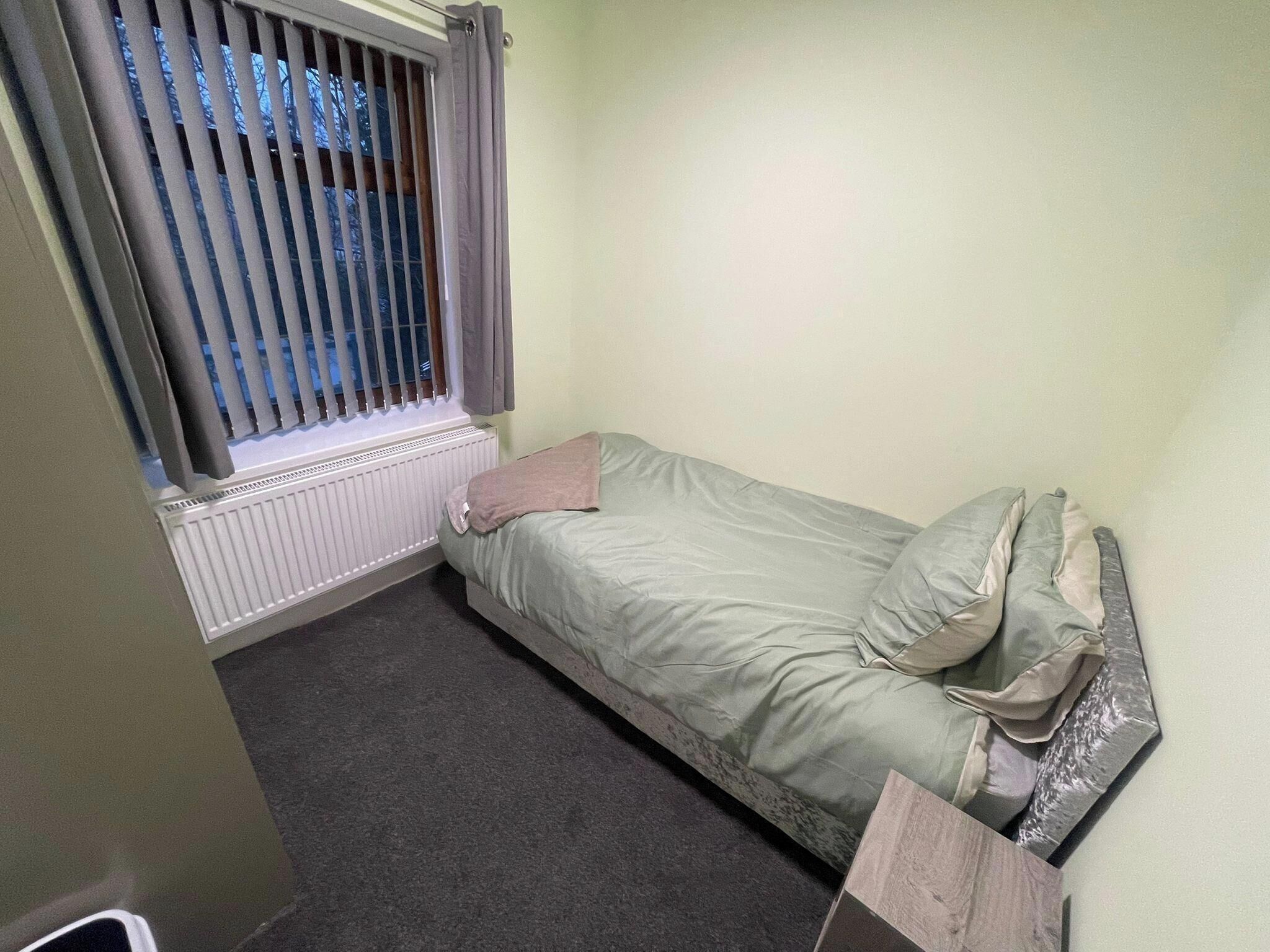 A small, single-bed room with a window, a radiator, and a wooden headboard.