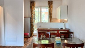 Ein Essbereich mit Tisch und Stühlen, ein Küchenbereich mit Kühlschrank und ein Fenster mit Vorhängen.