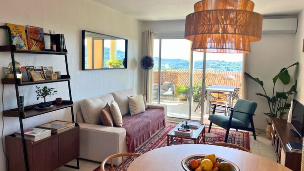Uma sala de estar com sofá, uma mesa de jantar com frutas, uma estante de livros e vista para o exterior.
