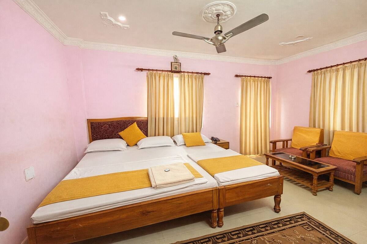 Une chambre d’hôtel avec un grand lit, un bureau, deux chaises, un ventilateur de plafond et des rideaux.