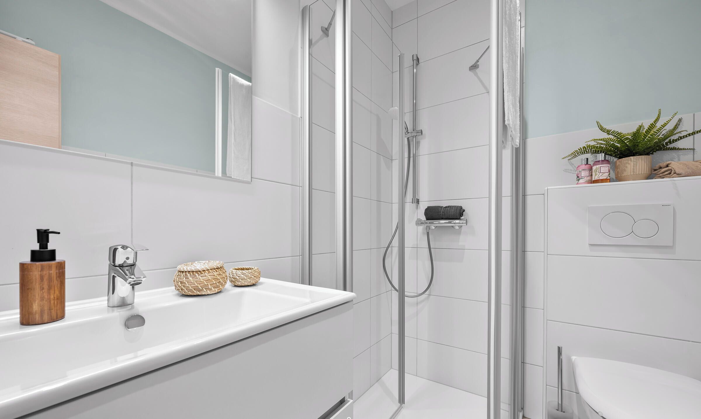 Un baño moderno con ducha envuelta en vidrio, un lavabo blanco con dispensador de jabón de madera y un espejo sobre el lavabo.
