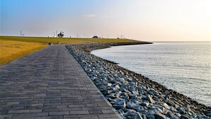 A paved walkway beside a rocky shoreline with a grassy area and distant structures. - “Apartment Amrum” in the heart of Büsum with balcony, centrally located and clos (Büsum)