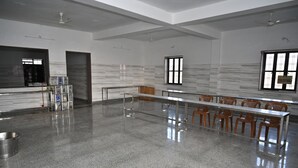 Um quarto amplo com uma mesa comprida e cadeiras, um ventilador de teto e uma janela com persianas.