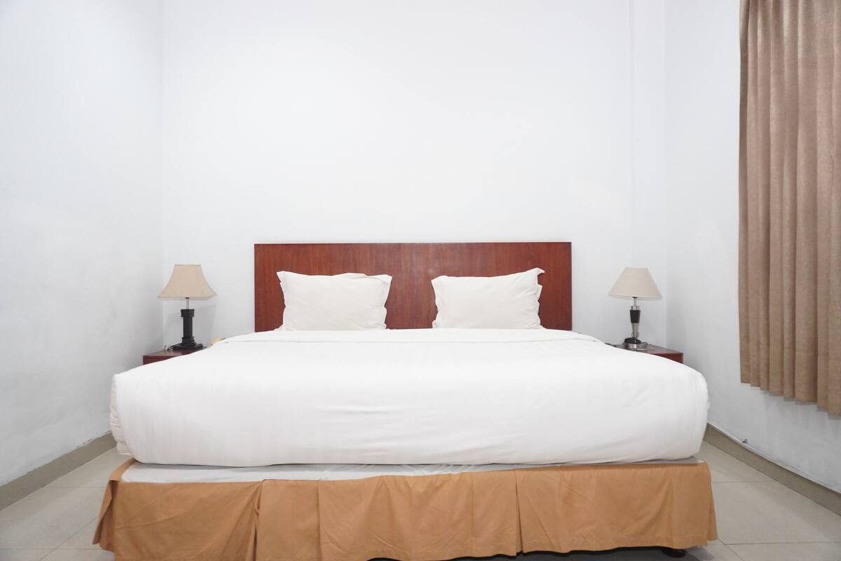 Tyypillinen hotellihuone, jossa on puinen pääty, kaksi valkoista tyynyä, ruskealla reunuksella varustettu sänky, kaksi yöpöytävalaisinta ja ikkuna, jossa on verhot.