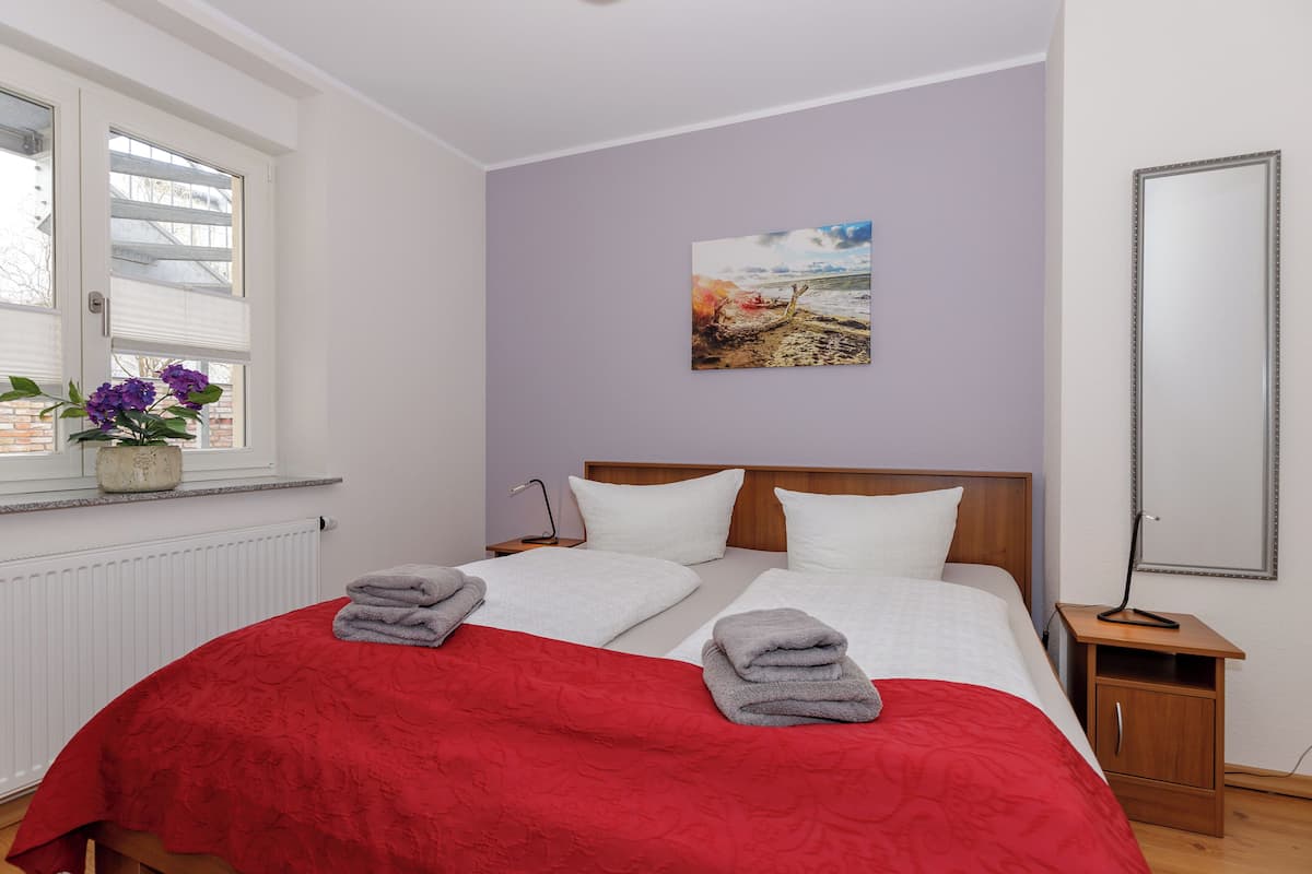 Un lit double avec une couvre-lit rouge, deux oreillers blancs et deux serviettes sur le lit. Il y a une fenêtre donnant sur un immeuble, et une plante est posée sur le rebord de la fenêtre. Un tableau est accroché au mur, et un miroir se trouve sur le mur, à droite.