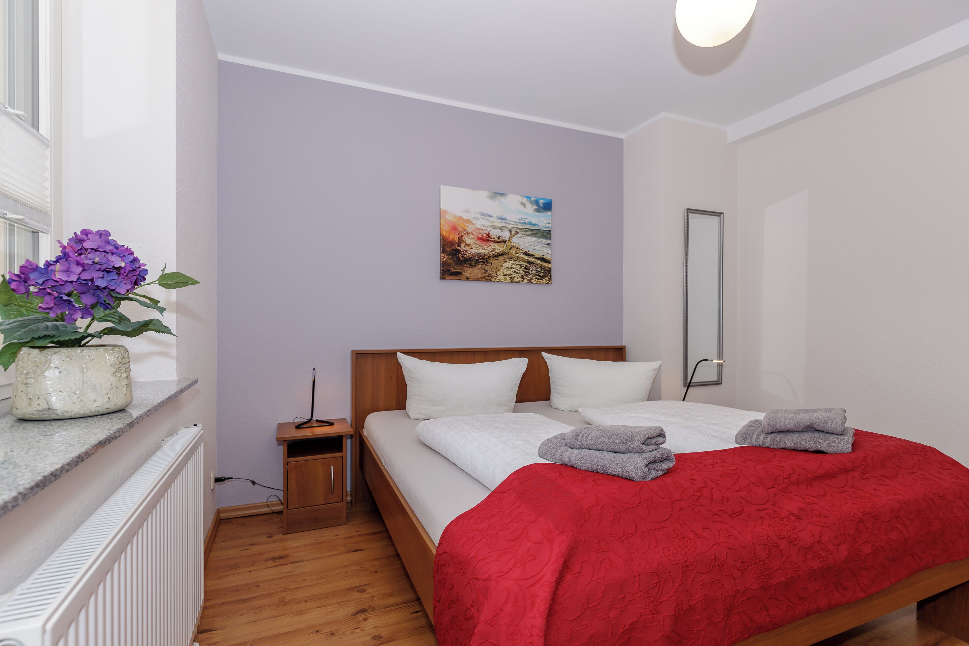 A bedroom with a bed, a nightstand, a bedside table, a potted plant, a painting on the wall, and a window.