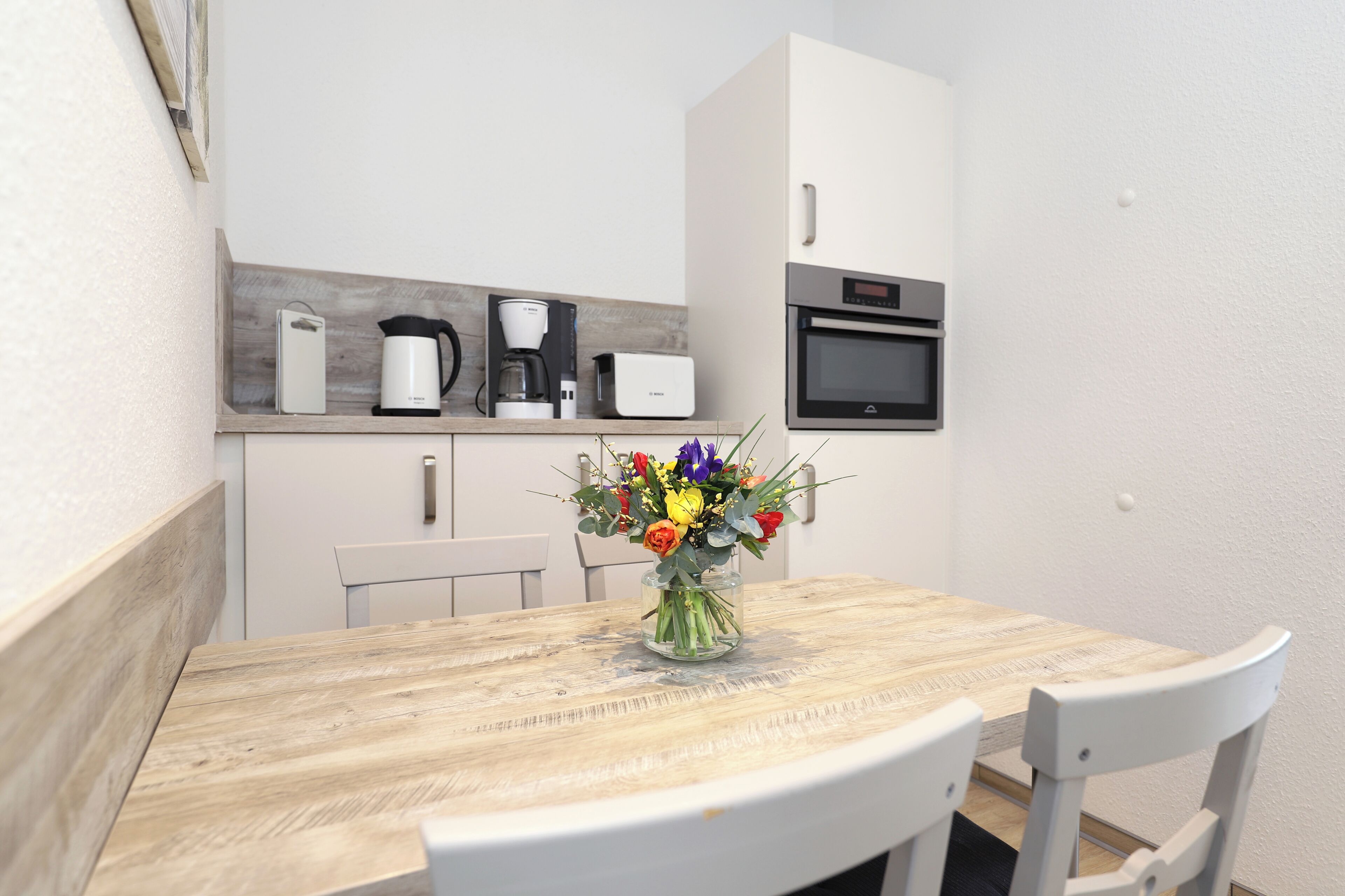 Eine moderne Küche mit Holztisch, weißen Stühlen, einem Blumenstrauß und einer Mikrowelle über der Arbeitsfläche.
