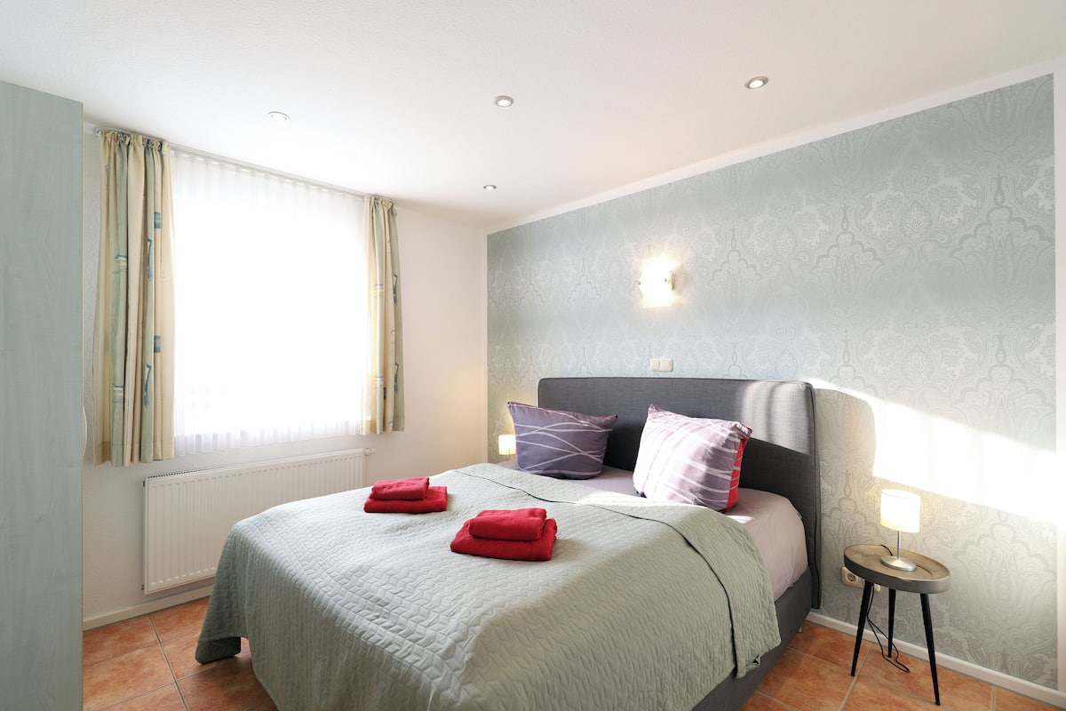 Une chambre à coucher avec un lit, des oreillers, une lampe de chevet, une fenêtre avec des rideaux et un radiateur.