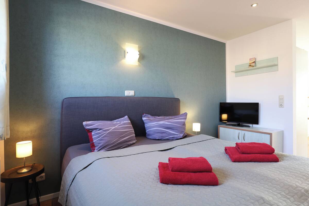 Une chambre d’hôtel équipée d’un lit, d’oreillers, d’une télévision, d’une table de chevet et d’une étagère fixée au mur.
