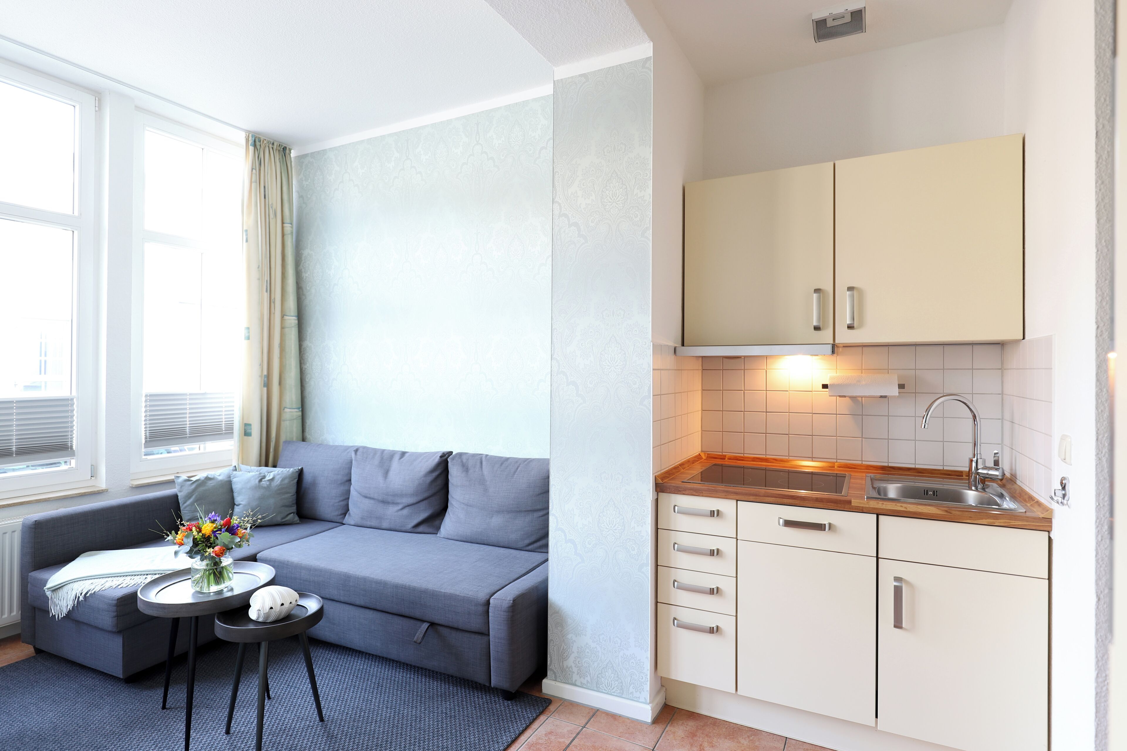 En kompakt opholdsstue med en sofa, et lille rundt bord med en vase blomster, et tekøkken med vask og skabe, samt et vindue med persienner.