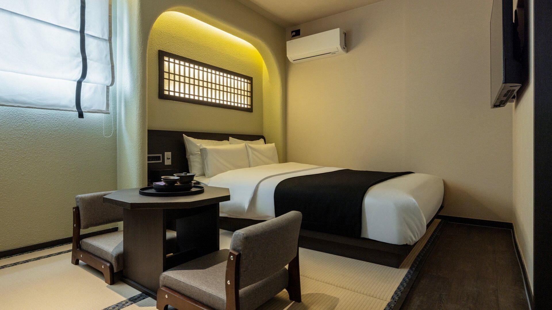 A hotel room with a bed, a small table with a tray, two chairs, a window with curtains, and an air conditioning unit.