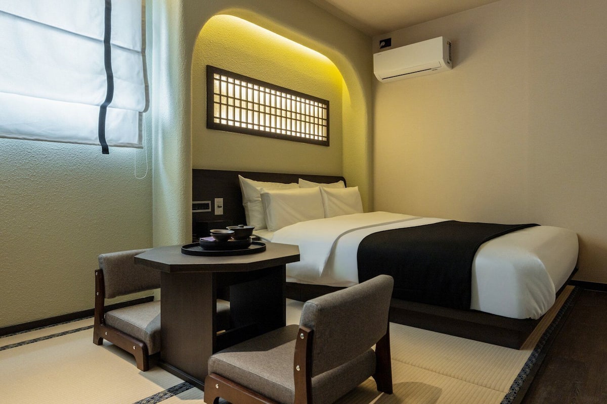 A hotel room with a bed, a small table with a tray, two chairs, a window with curtains, and an air conditioning unit.