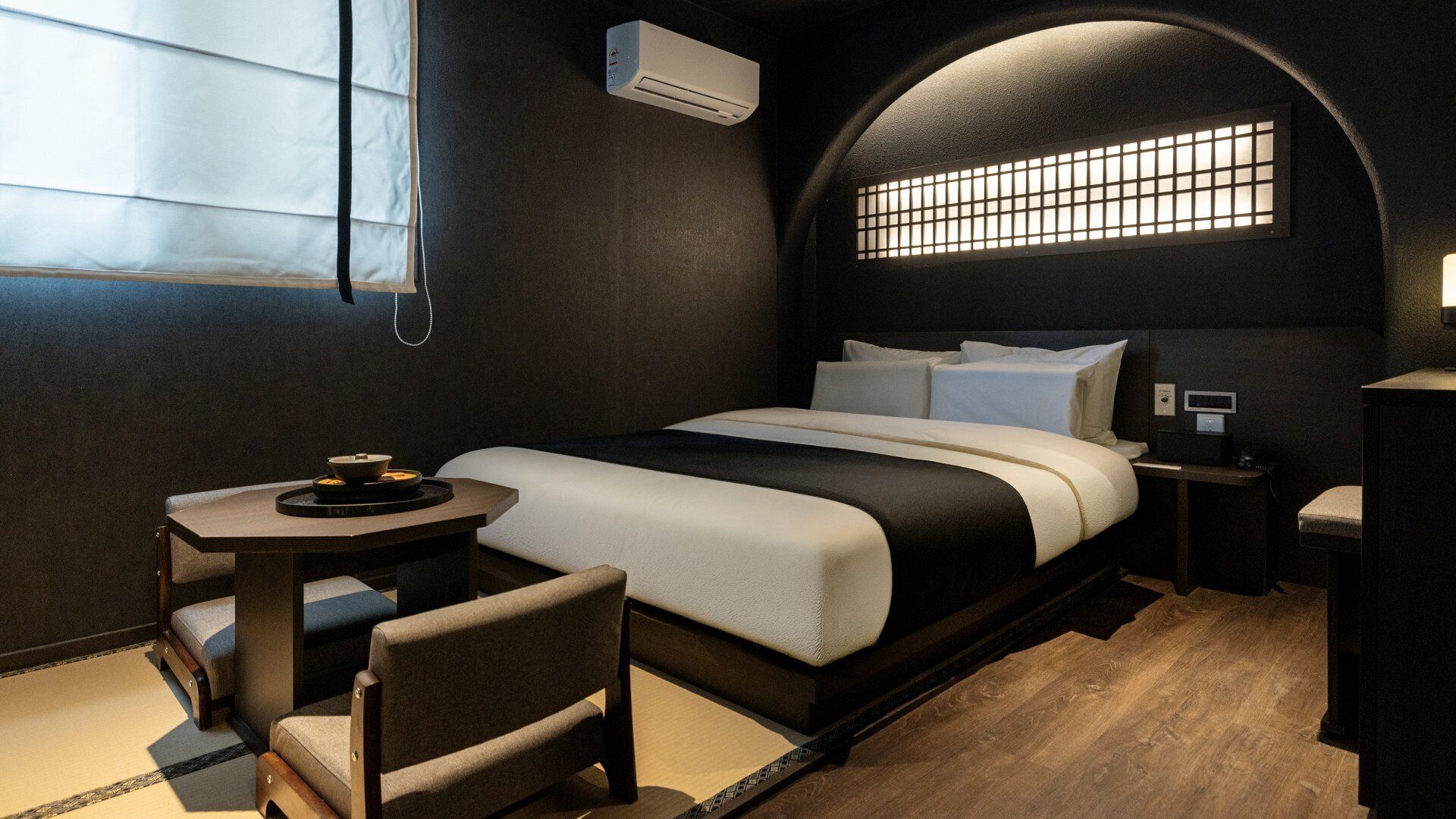A modern hotel room with a bed, bedside table, chair, and a small table with a bowl.