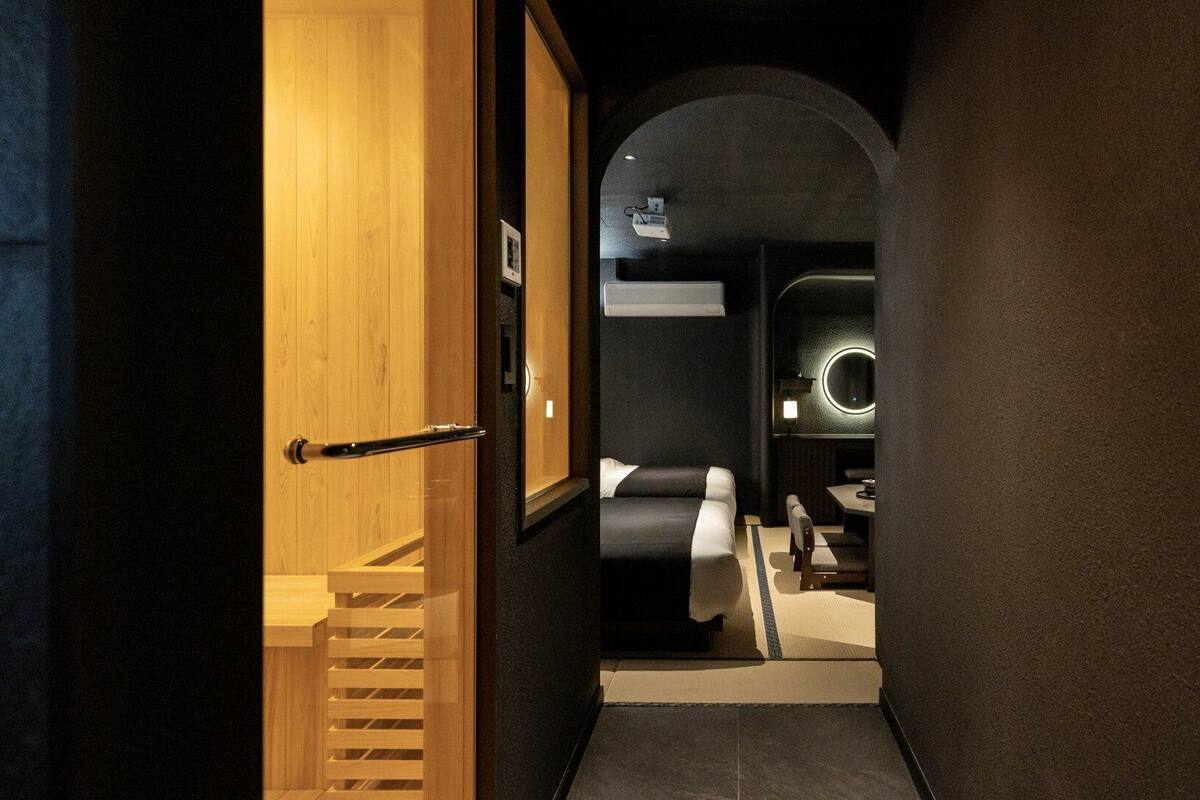 A hotel hallway with a wooden door, a staircase, a bed, and a bathroom with a circular mirror.