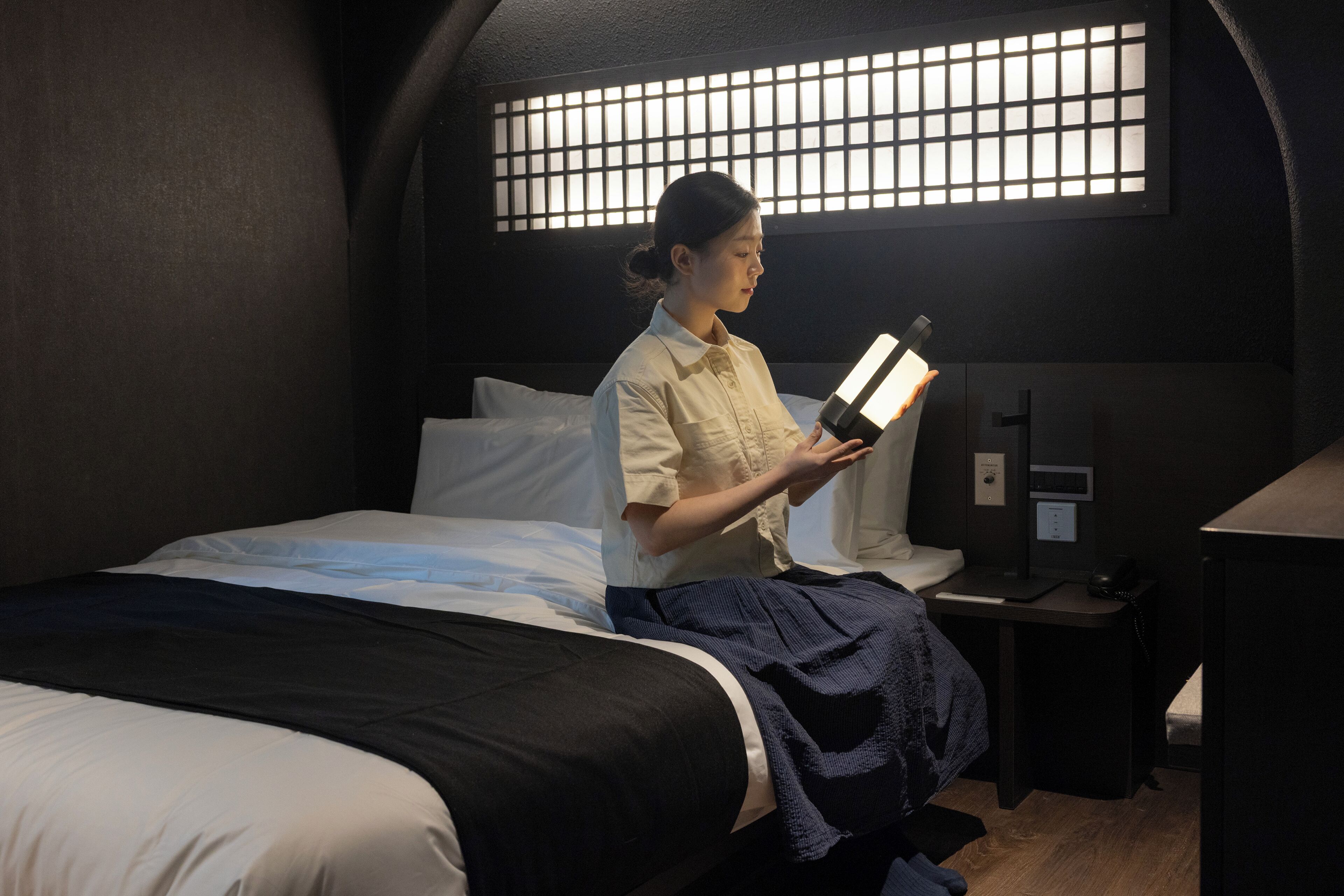 A room with a bed, a bedside table, a nightstand, and a window with a grid pattern.