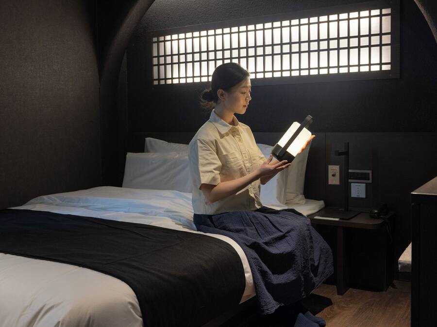 A room with a bed, a bedside table, a nightstand, and a window with a grid pattern.