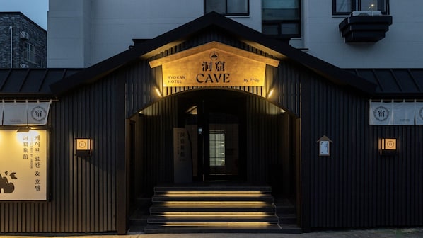 Hotellin sisäänkäynti, jonka julkisivu on tumma, ja jossa on kyltti, jossa lukee: »Ryokan Hotel Cave by Aonok», sekä portaat, jotka johtavat ovelle.