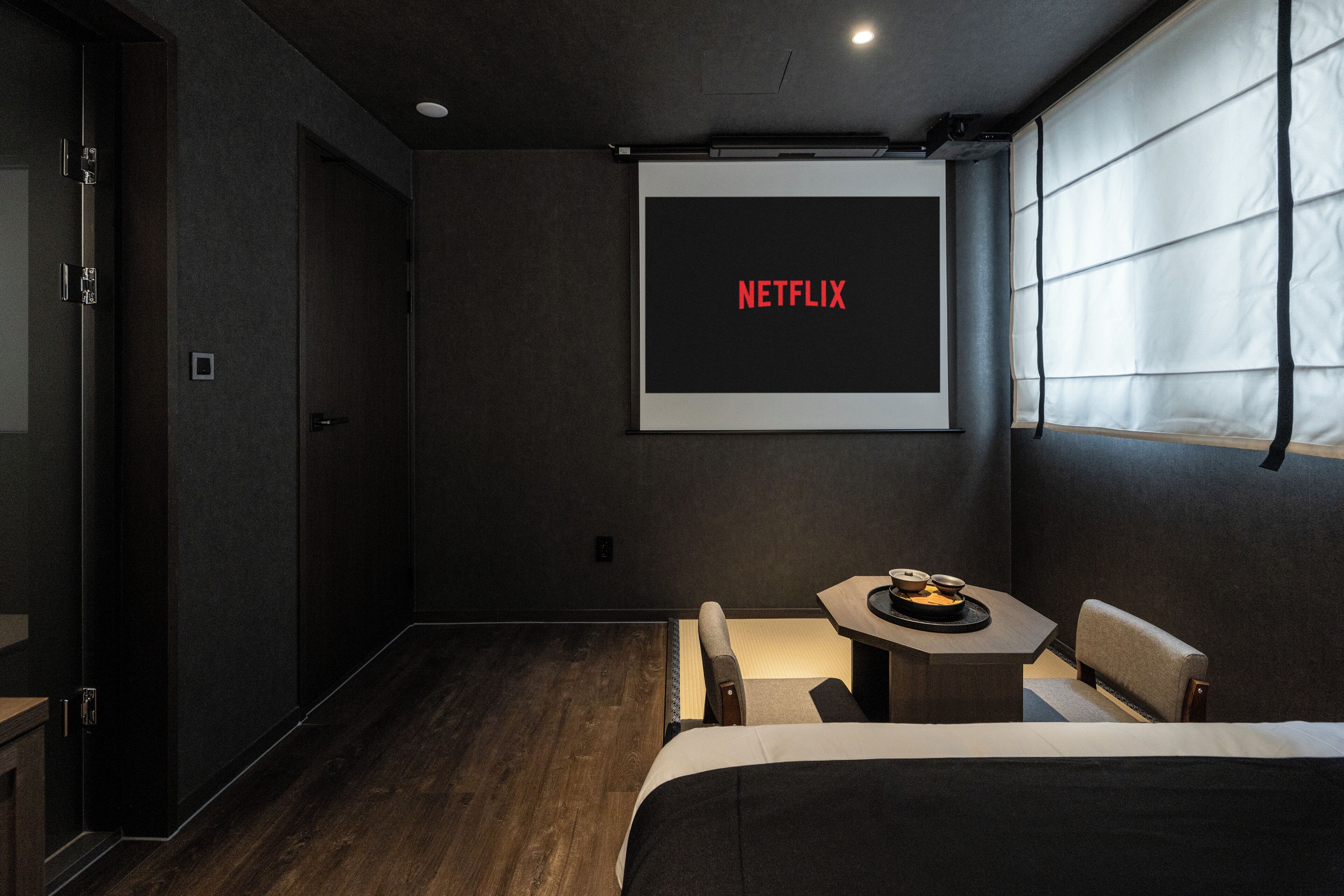 A modern hotel room with a bed, a small table with a tray of snacks, a wall-mounted screen displaying 'NETFLIX,' and a window with blinds.