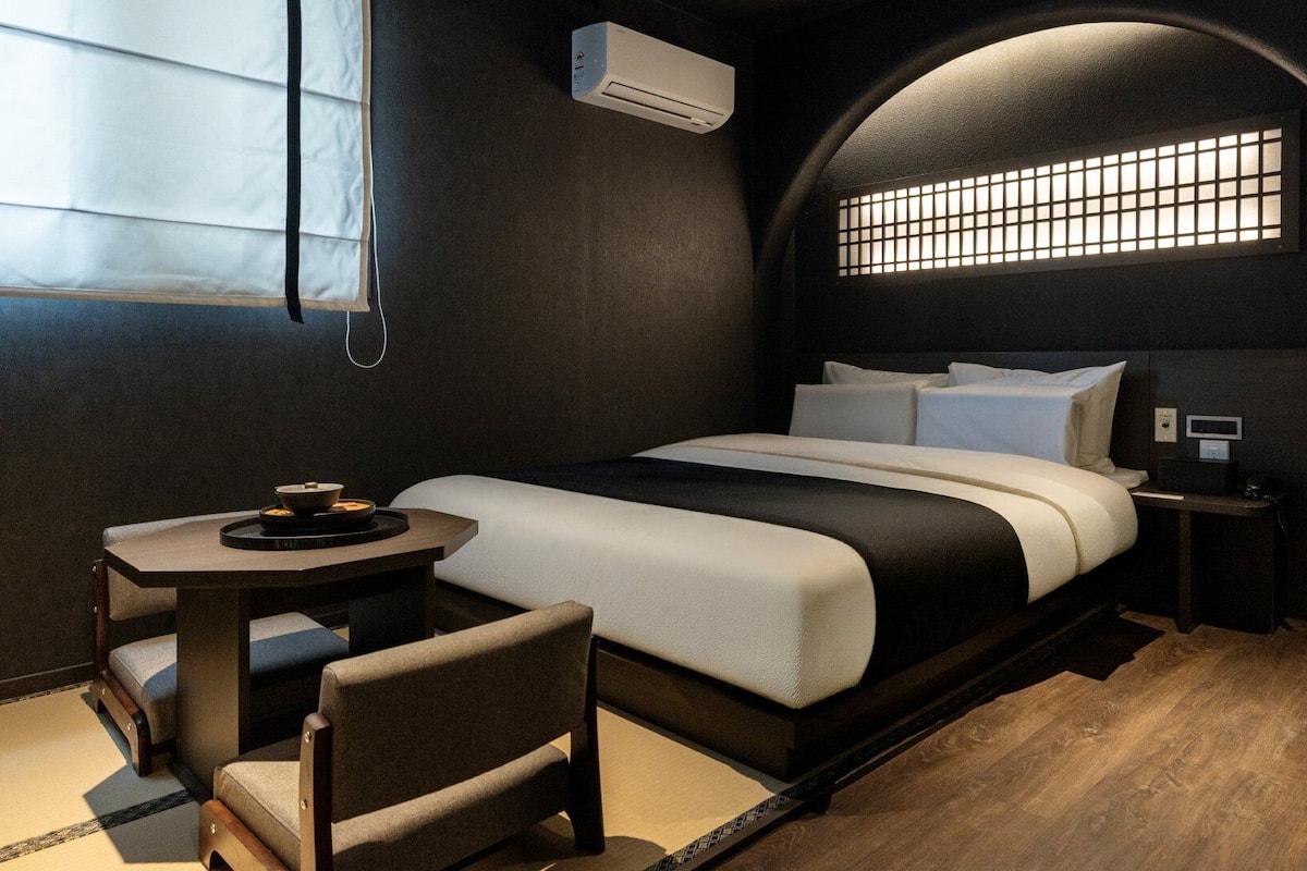 A modern hotel room with a bed, bedside table, chair, and a small table with a bowl.
