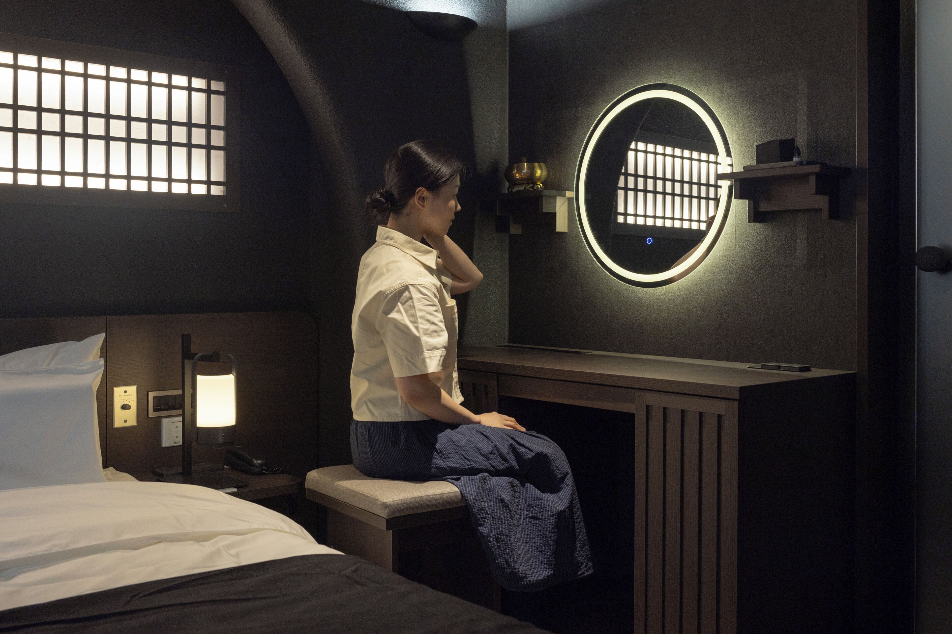 A hotel room with a bed, a desk with a chair, a round mirror, a nightstand, and a wall-mounted shelf.