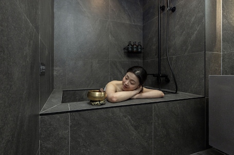 A person relaxing in a modern bathroom with a bathtub, a small shelf with bottles, and a sign indicating a shower area.. Cave Ryokan Hotel by Anook Daejeon Yucheon Southwest Terminal
