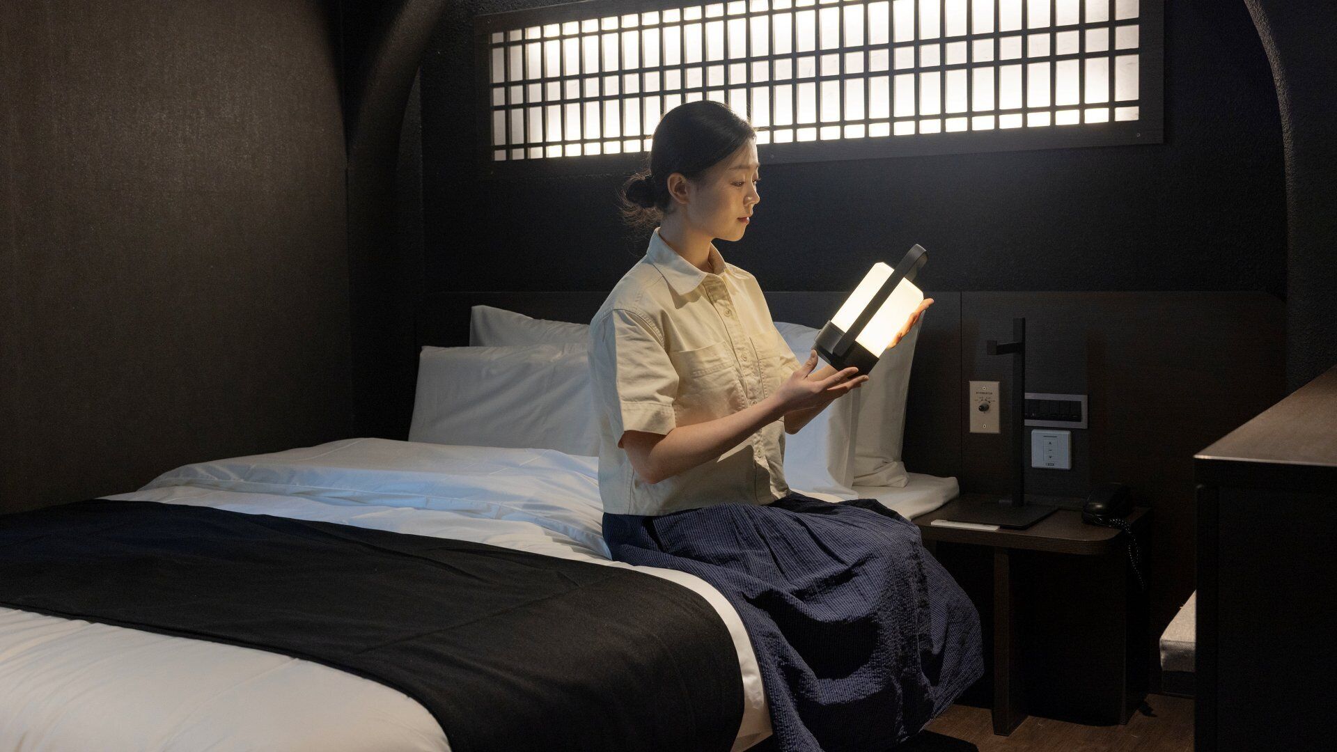 A person sitting on a bed in a room with a window, a bedside table, and a nightstand.