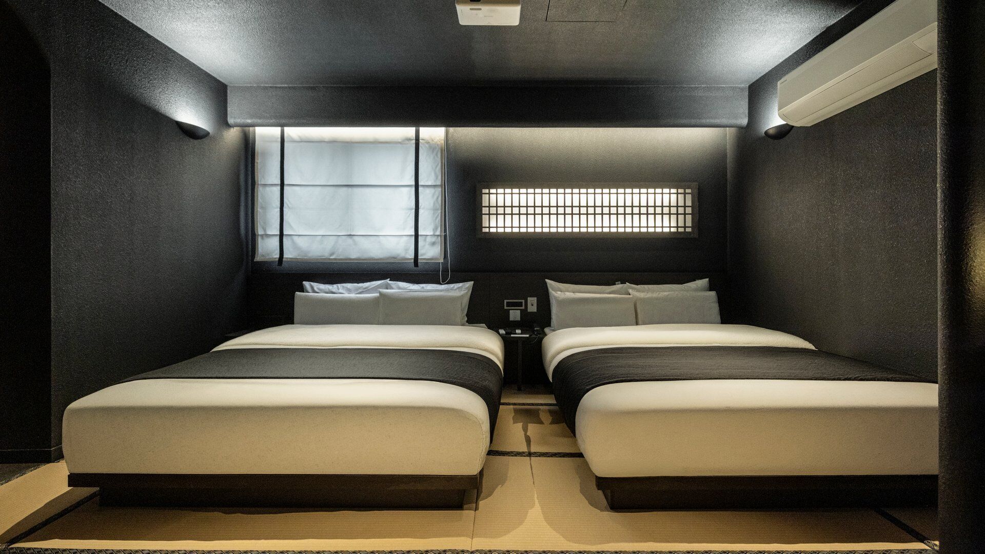 A small, dimly lit room with two beds, a window with blinds, and a small table with a phone.