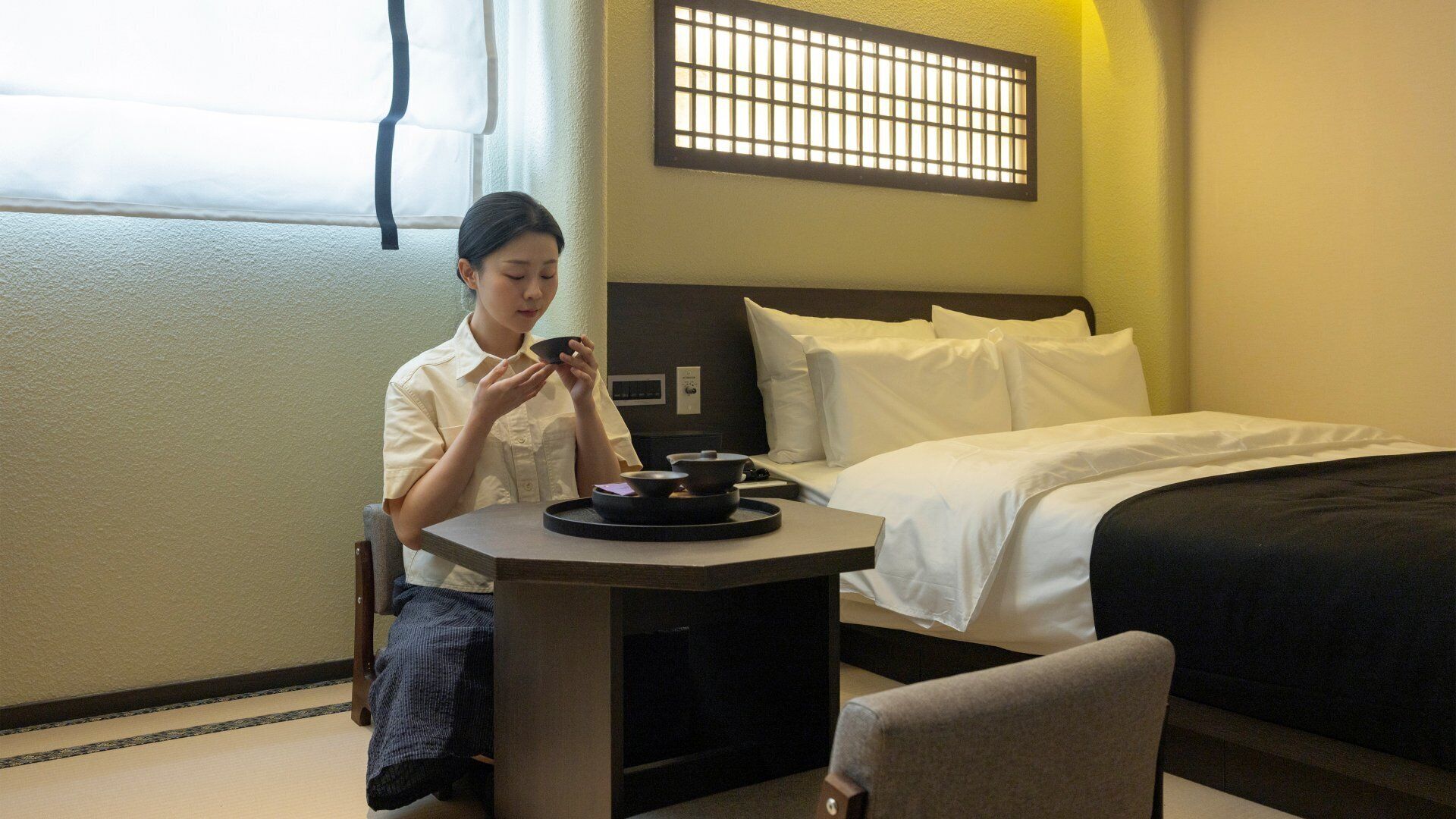 Une chambre d’hôtel avec un lit, une petite table sur laquelle se trouve un plateau avec divers objets, une chaise et une fenêtre avec des stores.