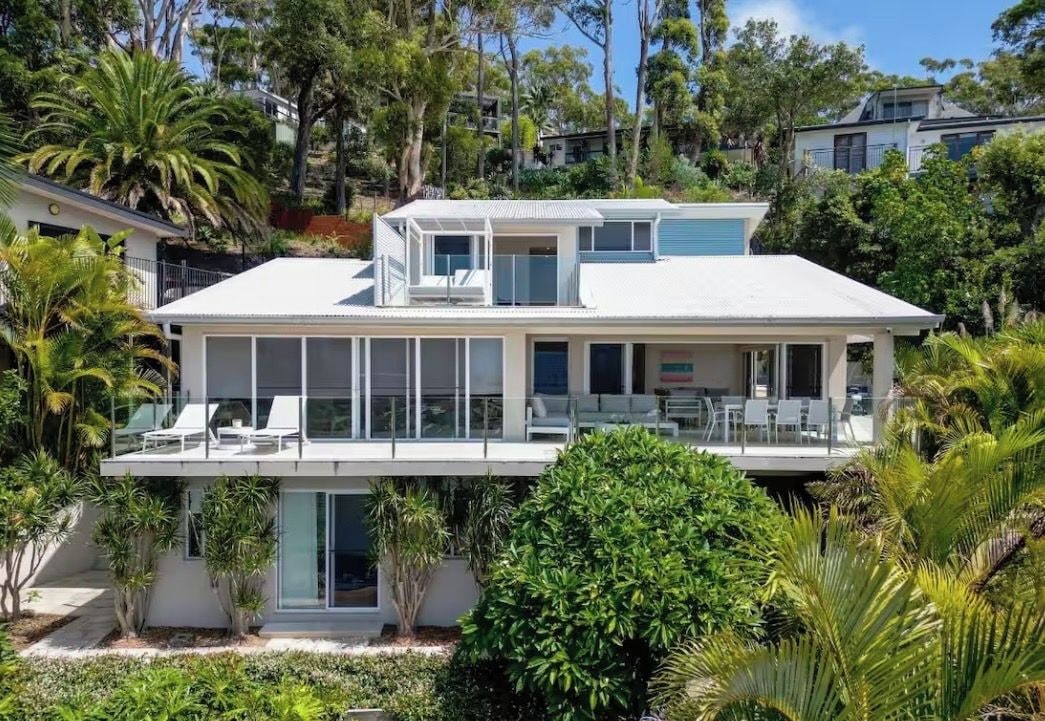A modern two-story house with a balcony, surrounded by lush greenery.